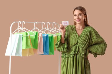 Güzel, genç, mutlu bir kadın hediye kartıyla ve bej arka planda raflarda alışveriş torbalarıyla