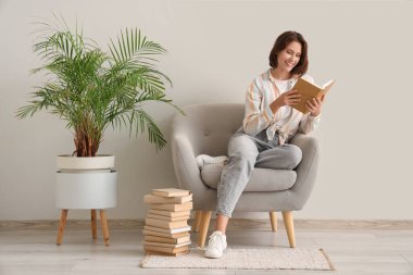 Beyaz duvarın yanındaki koltukta kitap okuyan güzel bir kadın.
