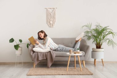 Beyaz duvarın yanındaki kanepede kitap okuyan güzel bir kadın.