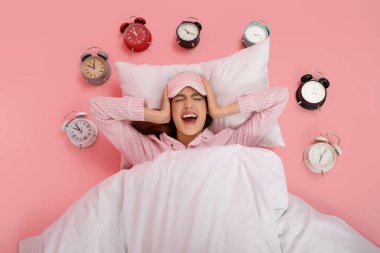 Güzel genç bir kadın çalar saatlerle yerde yatıyor ve pembe arka planda çığlık atıyor.