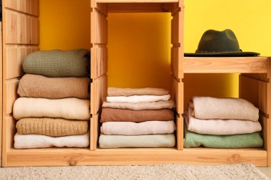 Farklı katlanmış kıyafetleri ve sarı duvarın yanında şapkası olan ahşap raf ünitesi, yakın plan.