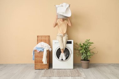 Çamaşır sepetiyle odada çamaşır makinesinde oturan genç bir kadın.