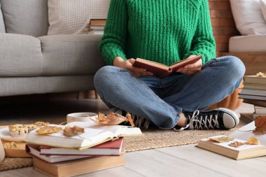 Evde halıda sonbahar yapraklarıyla kitap okuyan bir kadın.