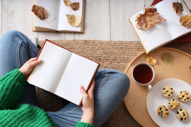 Boş kitabı ve sonbahar yaprakları olan bir kadın evde, halıda oturuyor.