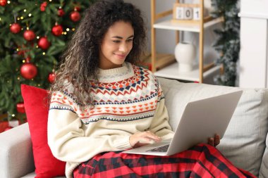 Güzel, mutlu, Afro-Amerikan bir kadın dizüstü bilgisayarıyla Noel için dekore edilmiş bir odada oturuyor.
