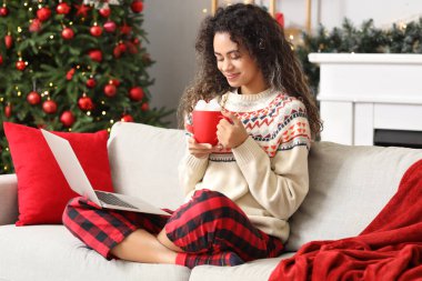 Güzel, mutlu, Afro-Amerikan bir kadın, elinde bir fincan kakao ve dizüstü bilgisayarla Noel için dekore edilmiş bir odada, kanepede oturuyor.