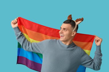 Mavi arka planda LGBT bayrağı olan ren geyiği boynuzlu genç bir adam.