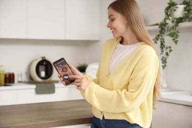 Mutfakta akıllı ev güvenlik kontrol paneli olan cep telefonu kullanan genç bir kadın.