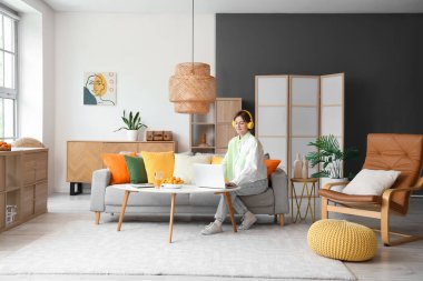 Oturma odasında kanepede otururken dizüstü bilgisayar kullanan kulaklıklı genç bir kadın.