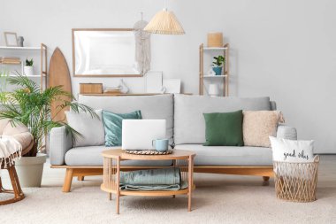 Aydınlık oturma odasının içinde gri kanepe ve sehpanın üzerinde modern dizüstü bilgisayar.