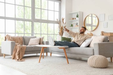 Uzaktan kumandalı genç adam evdeki rahat koltukta televizyon izliyor.