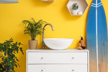 Modern lavabo, çekmeceler ve banyonun içindeki sarı duvarın yanında sörf tahtası.