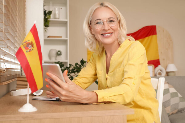 Mature woman with tablet computer and flag of Spain learning language online at table