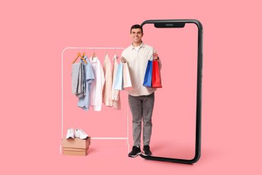 Büyük bir cep telefonu ve pembe arka planda asılı şık giysileri olan genç bir adam.