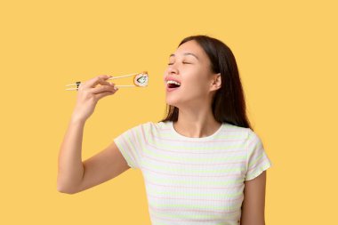 Güzel bir Asyalı kadın sarı arka planda çubuklarla lezzetli suşi yiyor.