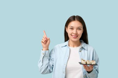 Enfes suşi tepsileriyle genç ve güzel bir Asyalı kadın mavi arka plandaki bir şeyi işaret ediyor.