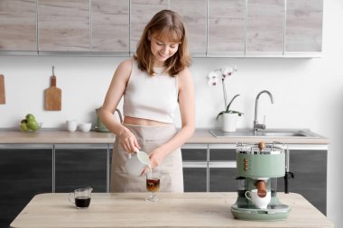 Mutfakta buzlu kahveyle sürahiden bardağa süt döken genç bir kadın.