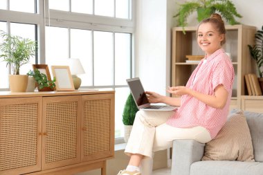 Evdeki koltukta modern laptop kullanan genç bir kadın.
