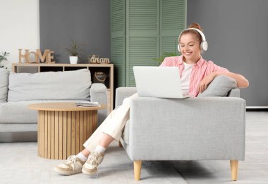 Kulaklıklı genç bir kadın evdeki koltukta modern laptop kullanıyor.