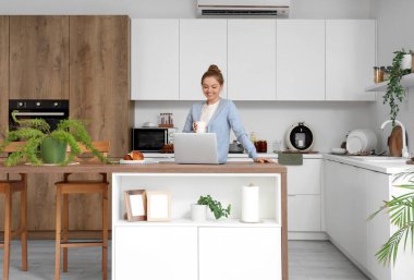 Mutfaktaki masada bir fincan kahve ve modern dizüstü bilgisayarı olan genç bir kadın.