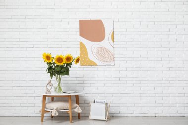 Oturma odasındaki beyaz tuğla duvarın yanındaki masada güzel ayçiçekleri olan heykel, dergi ve vazo.