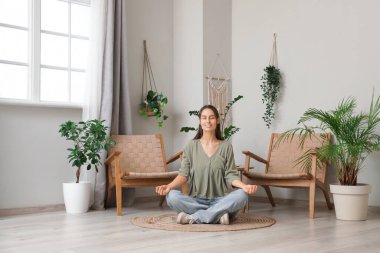 Genç bir kadın evde yerde oturmuş meditasyon yapıyor.