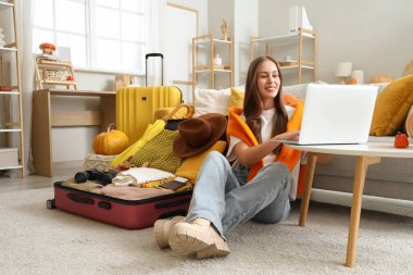 Sonbahar günü evde dizüstü bilgisayar kullanan kadın turist bavullarını topluyor.