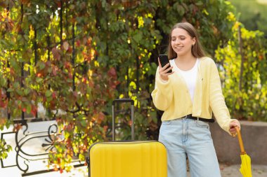 Şemsiyeli ve çantalı kadın turist, sonbahar günü parkta cep telefonu kullanıyor.