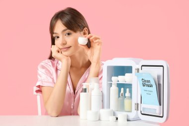 Pembe arka planda kozmetik ürünleri için küçük buzdolabı olan güzel genç bir kadın.