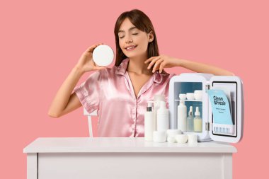 Pembe arka planda kozmetik ürünleri için küçük buzdolabı olan güzel genç bir kadın.
