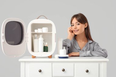 Gri arka planda kozmetik ürünleri için küçük buzdolabı olan güzel genç bir kadın.