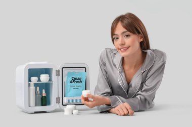 Gri arka planda kozmetik ürünleri için küçük buzdolabı olan güzel genç bir kadın.