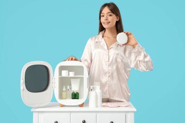 Mavi arka planda kozmetik ürünleri için küçük buzdolabı olan güzel genç bir kadın.