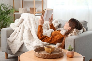 Evdeki kanepede sıcak battaniye okuyan genç bir kadın.