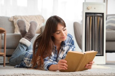 Evde elektrikli ısıtıcının yanında kitap okuyan genç bir kadın.