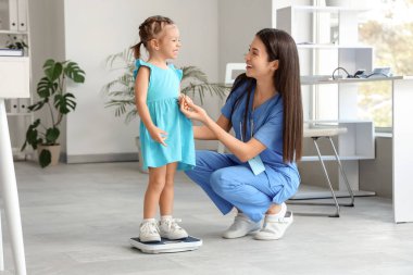 Klinikte tartıya ağırlığını koyan küçük tatlı bir kızı olan kadın bir çocuk doktoru.
