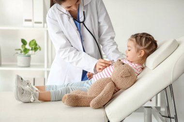 Steteskoplu kadın bir çocuk doktoru klinikteki kanepede tatlı küçük bir kızı dinliyor.