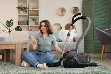 Cep telefonu ve elektrikli süpürgeyle oturma odasında halının üzerinde oturan genç bir kadın.