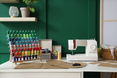 Atölyedeki yeşil duvarın yanındaki terzi masasında desenli dikiş makinesi ve iplik makaraları var.