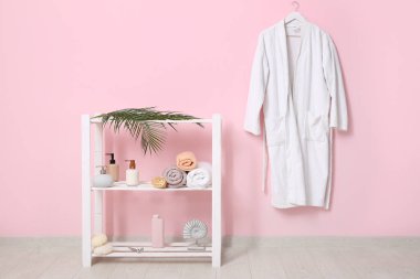 Pembe duvarda asılı beyaz bornoz ve banyoda raflarda farklı banyo malzemeleri.