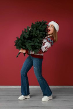 Kırmızı arka planda Noel ağacı olan Noel Baba şapkalı genç bir kadın.