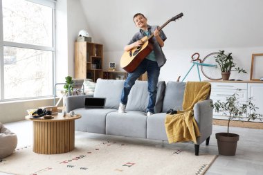 Genç çocuk oturma odasındaki kanepede gitar çalıyor.