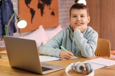 Genç çocuk dizüstü bilgisayarla masada oturuyor ve odada ödev yapıyor.