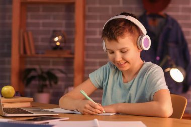 Kulaklıklı genç çocuk masada oturuyor ve odada ödev yapıyor.