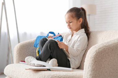 Sırt çantası, defter ve kalemle kanepede oturup evde ders çalışan sevimli küçük bir kız.