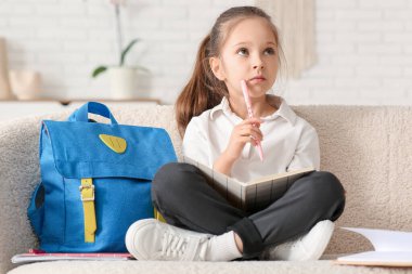 Sırt çantası, defteri ve kalemi olan düşünceli küçük kız kanepede oturup evde ders çalışıyor.