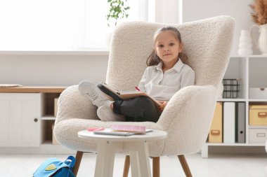 Not defteri ve kalemi olan sevimli küçük kız koltukta oturuyor ve evde ders çalışıyor.