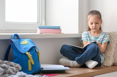 Sırt çantası, not defteri ve kalemle pencerenin yanında oturan ve evde ders çalışan sevimli küçük kız.