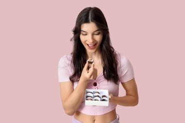 Pembe arka planda bir kutu lezzetli çikolata şekeri olan mutlu genç bir kadın.