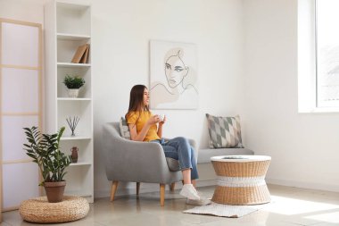 Genç bir kadın odada koltukta otururken bir fincan çay içiyor.